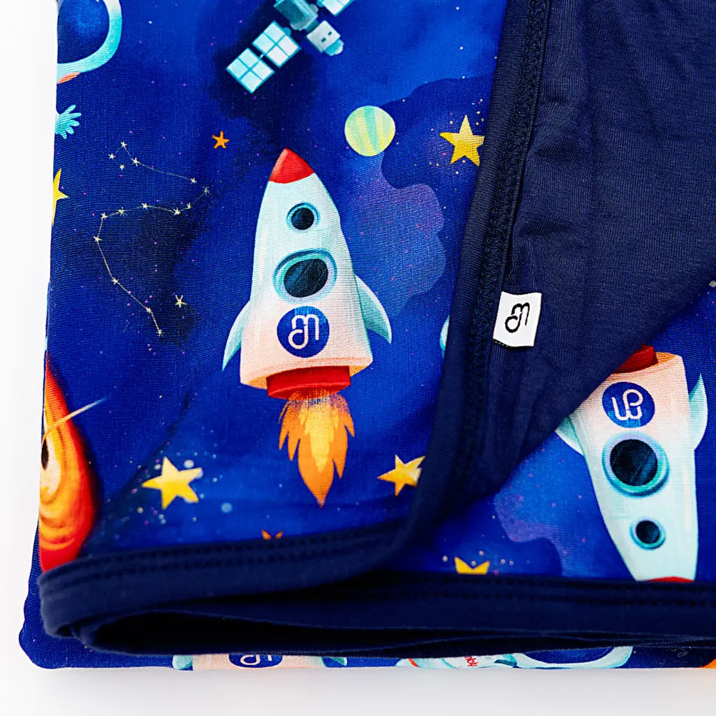 Close-up of a folded blanket with a royal blue space print of rockets and stars on one side and solid navy on the other.